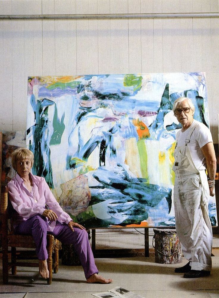 a man and woman standing in front of a large painting on easel next to each other