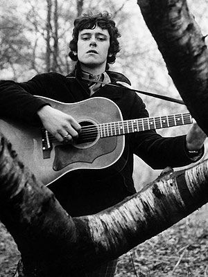 black and white photograph of man playing guitar in the woods with tree branches behind him