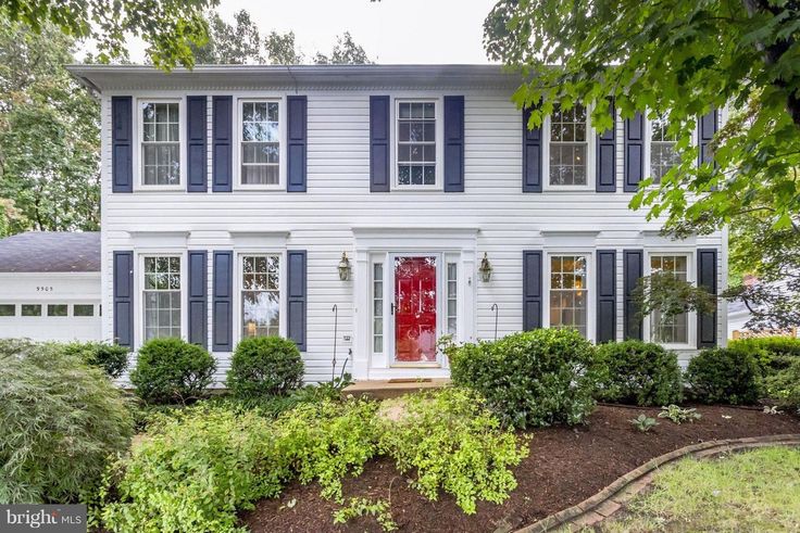 a white house with blue shutters on the front and red door, surrounded by trees