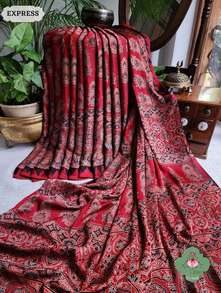 Update! - Zynah covered by LBB - https://lbb.in/bangalore/zynah-designs-handloom-sarees/ Collection: Daastaan-e-Ajrakh Transform your style with our Madder Red Ajrakh Modal Silk Saree. Immerse yourself in the rich heritage of hand block printing and eco-conscious fashion. Crafted with 100% natural dyes, this saree is a masterpiece of tradition and sustainability. Make a statement with elegance. Shop now for an exquisite addition to your wardrobe. About The Craft: Ajrakh Craft, rooted in Gujarat and Rajasthan, India, is a heritage-rich textile tradition celebrated for its intricate hand block printing and 100% natural dyes. Derived from "Azrak," meaning blue, the craft boasts a stunning range of colors and symmetrical patterns. Artisans meticulously carve wooden blocks to create detailed de Red Kalamkari Print Traditional Wear In Cotton Silk, Red Cotton Silk Traditional Wear With Kalamkari Print, Red Cotton Silk Blouse Piece With Kalamkari Print, Semi-stitched Red Saree With Block Print, Red Semi-stitched Block Print Saree, Red Semi-stitched Saree With Block Print, Red Block Print Semi-stitched Saree, Red Bohemian Blouse Piece With Bandhani Print, Red Block Print Mulmul Dupatta