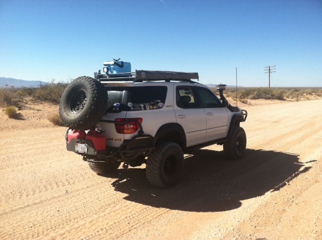 a white truck with large tires on the back driving down a dirt road in the desert