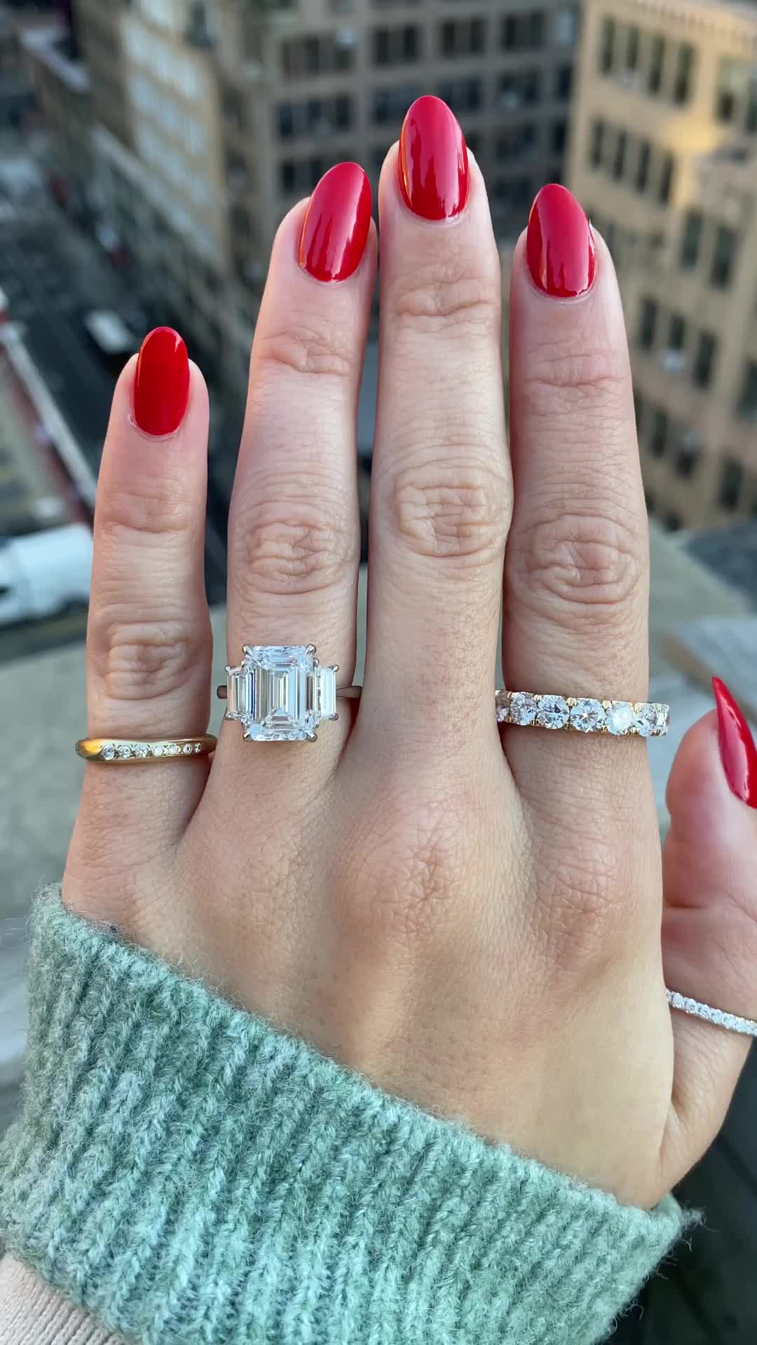 This may contain: a woman's hand with red fingernails and two rings on it, in front of a cityscape