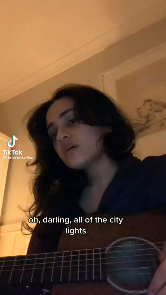 This may contain: a woman is playing an acoustic guitar in her living room with the caption'oh, daring all of the city lights '