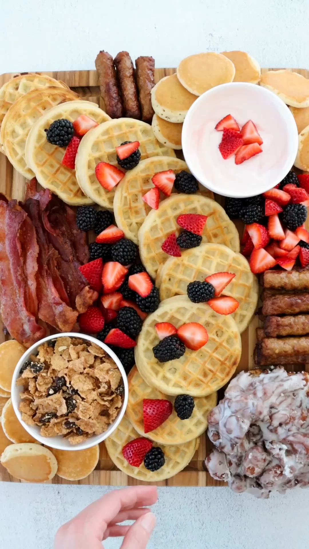 This contains: Waffles, pancakes, bacon, sausage, and fresh fruit arranged on a cheese board.