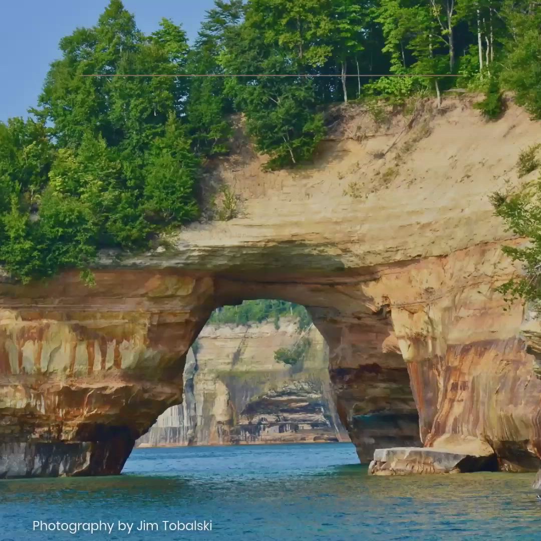 This may contain: an arch in the side of a cliff with trees on top and water below it