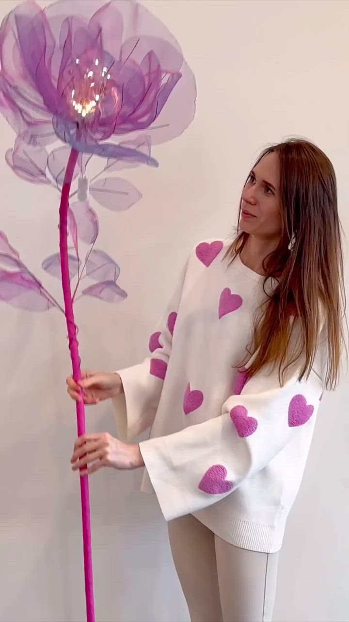 This may contain: a woman standing next to a purple flower with pink hearts on it's petals