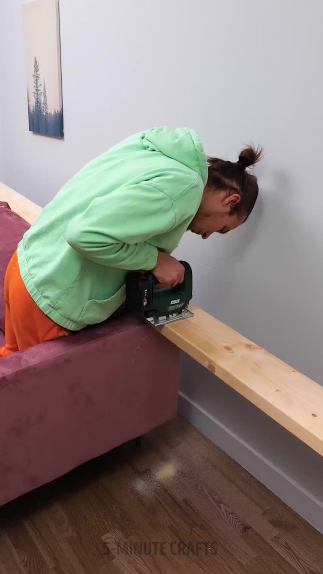 This may contain: a woman in green jacket sanding on bench with power driller and wood planks