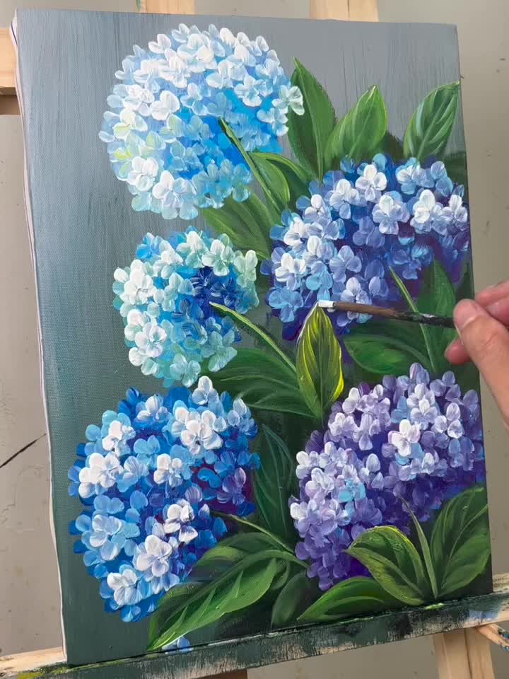 This may contain: a painting of blue and white flowers is being worked on by someone holding a paintbrush