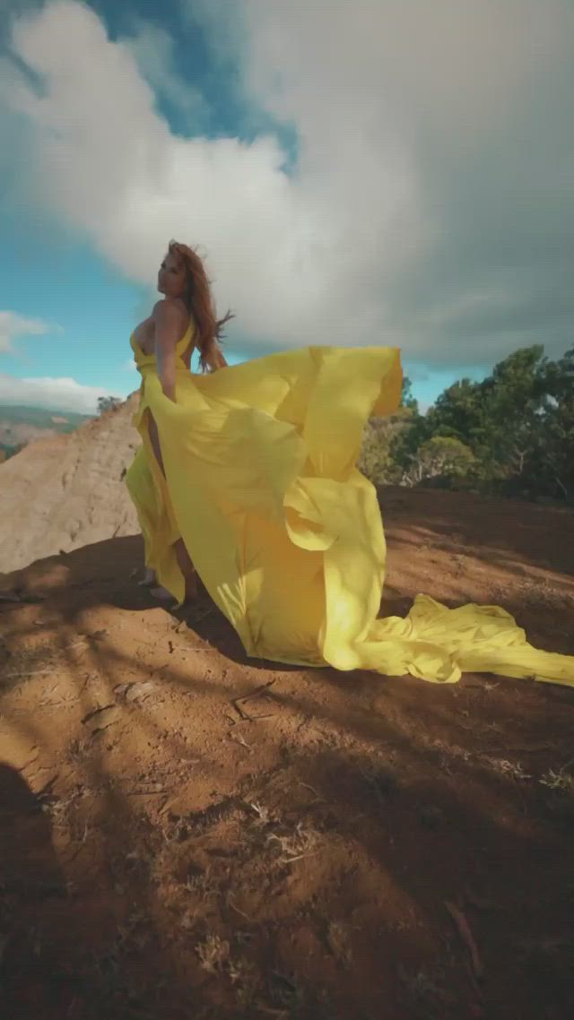 This may contain: a woman in a yellow dress sitting on top of a dirt hill next to trees