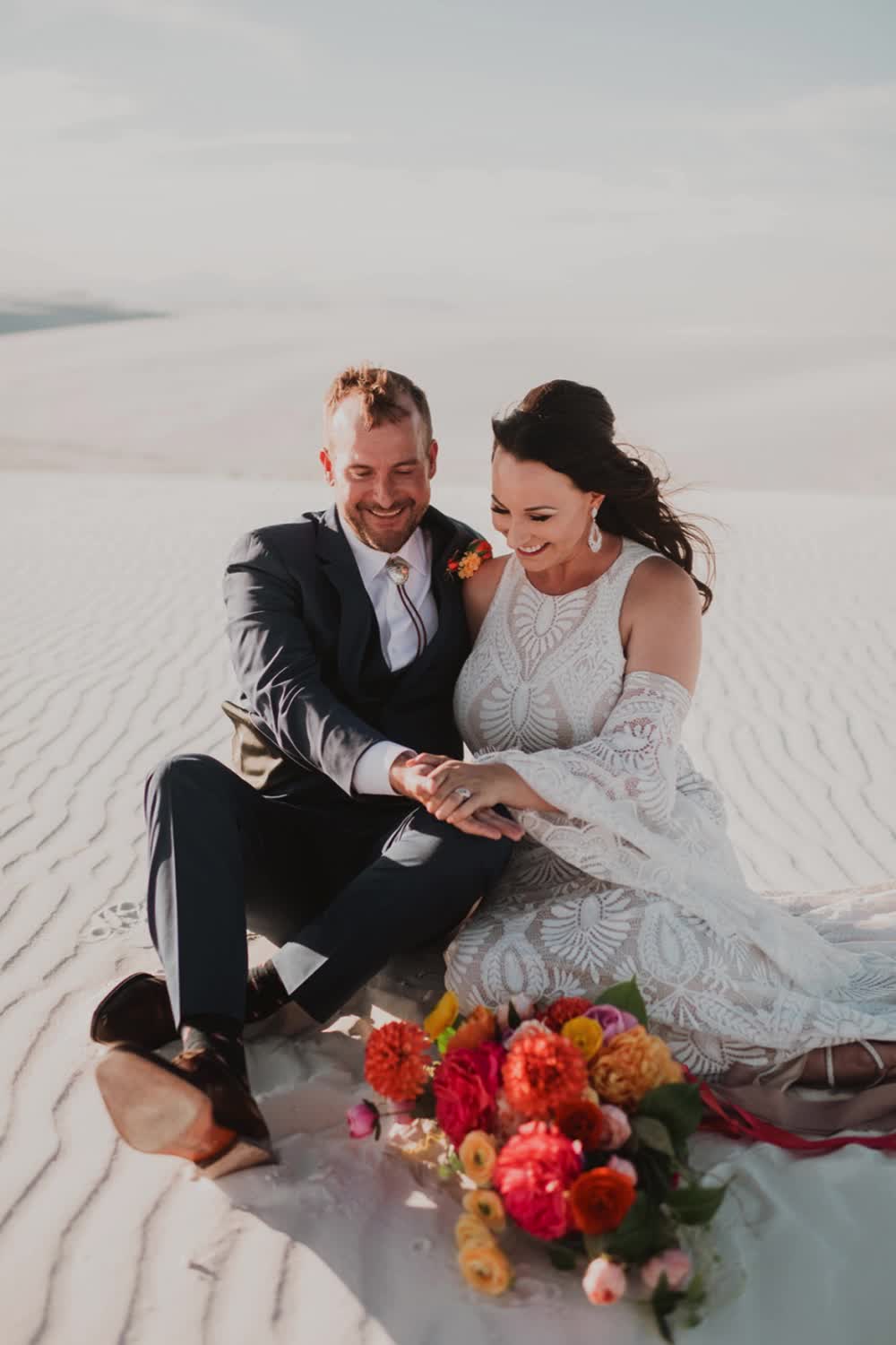 This may contain: a man and woman sitting on the sand with flowers in front of them, text reads make your wedding unforgettable with the enchanting marlowe dress