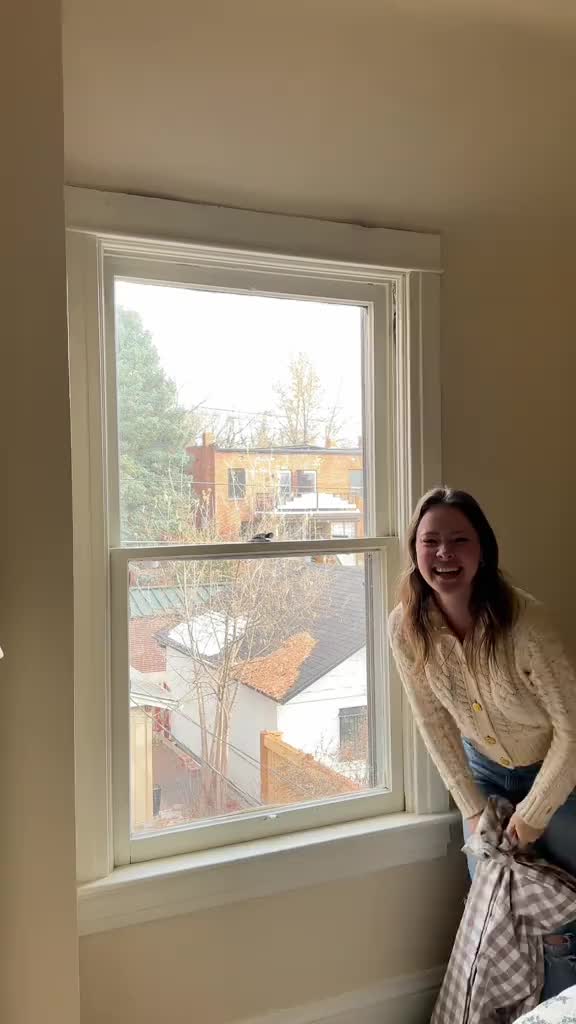 This may contain: a woman standing in front of a window with a blanket over her shoulder and smiling at the camera