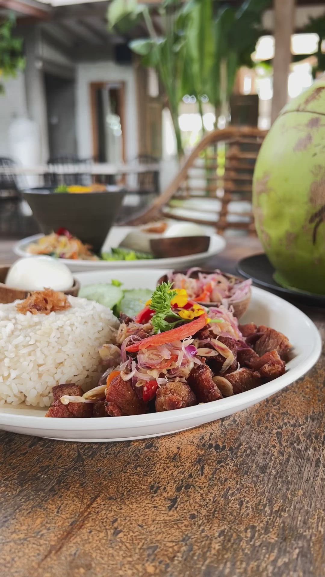 This may contain: a white plate topped with meat and rice next to a green coconut ball on top of a wooden table