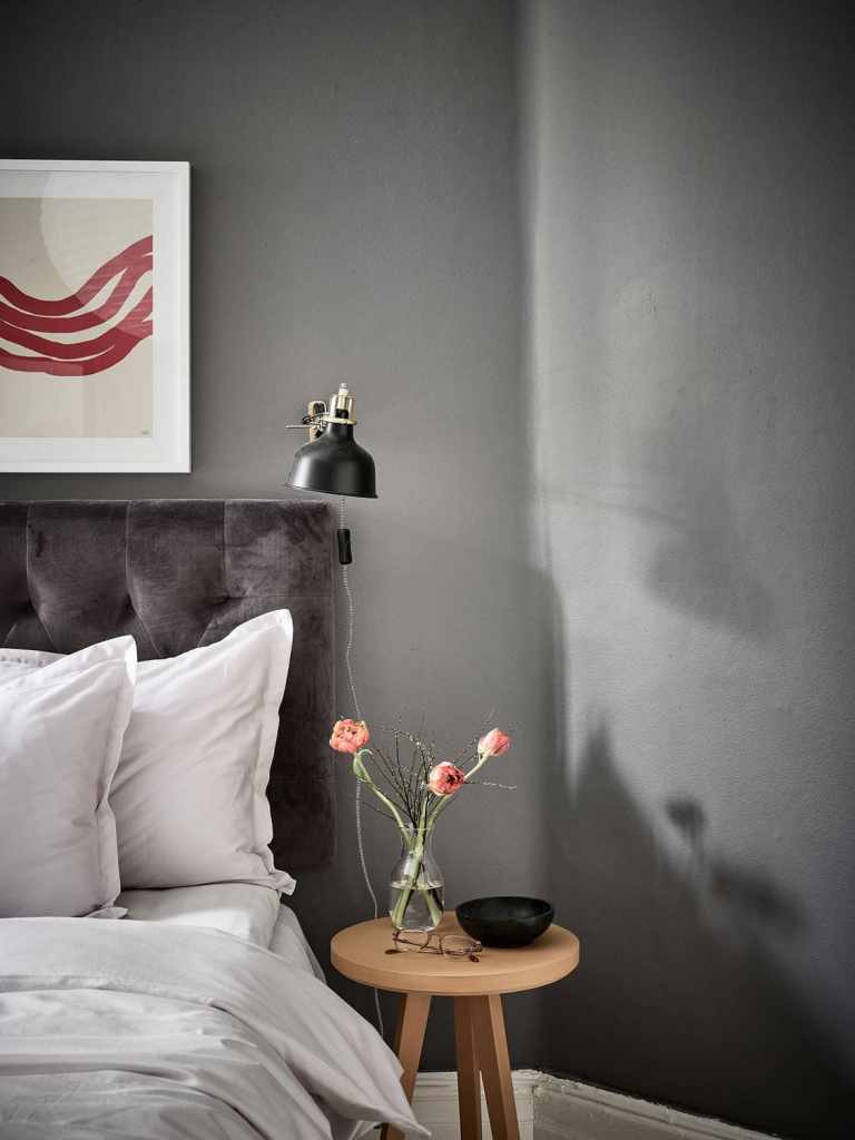 A bedroom with dark grey walls, a grey velvet headboard and white and beige bedding