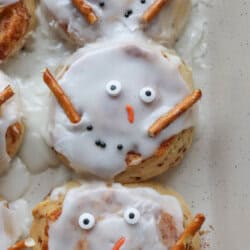 Cinnamon rolls decorated as snowmen, with white icing, candy eyes, pretzel arms, and orange candy noses, arranged in a baking pan.