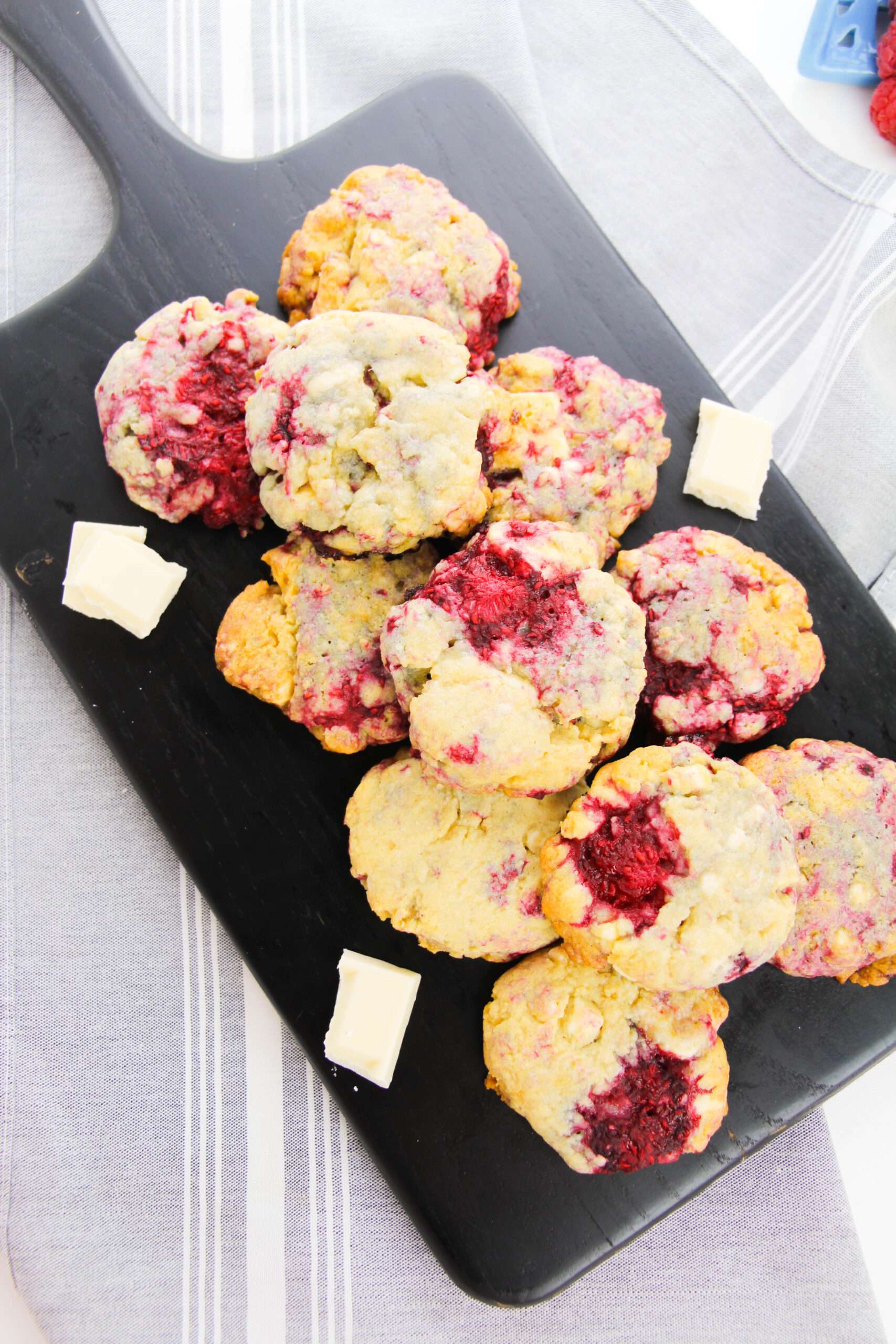 A black cutting board displays freshly baked Easy White Chocolate Raspberry Cookies with visible raspberry chunks and surrounded by small cubes of white chocolate. The board rests on a striped cloth.