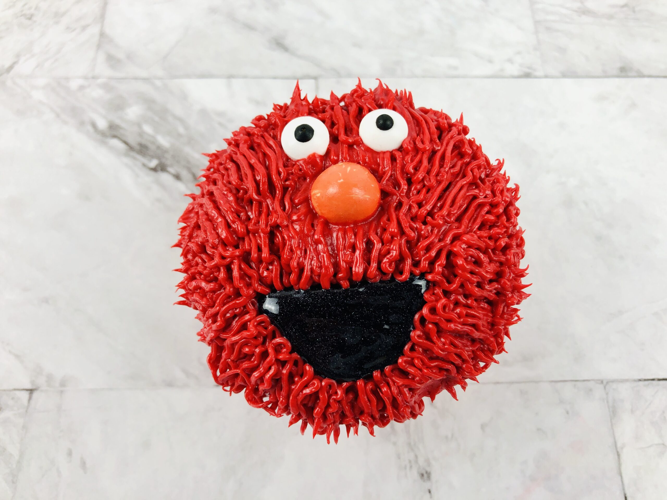 A cupcake decorated in red to resemble Elmo, with large white eyes and a round orange nose. The texture and color of the frosting create a playful and expressive appearance, with an open mouth made of black frosting. The background is a marble surface.