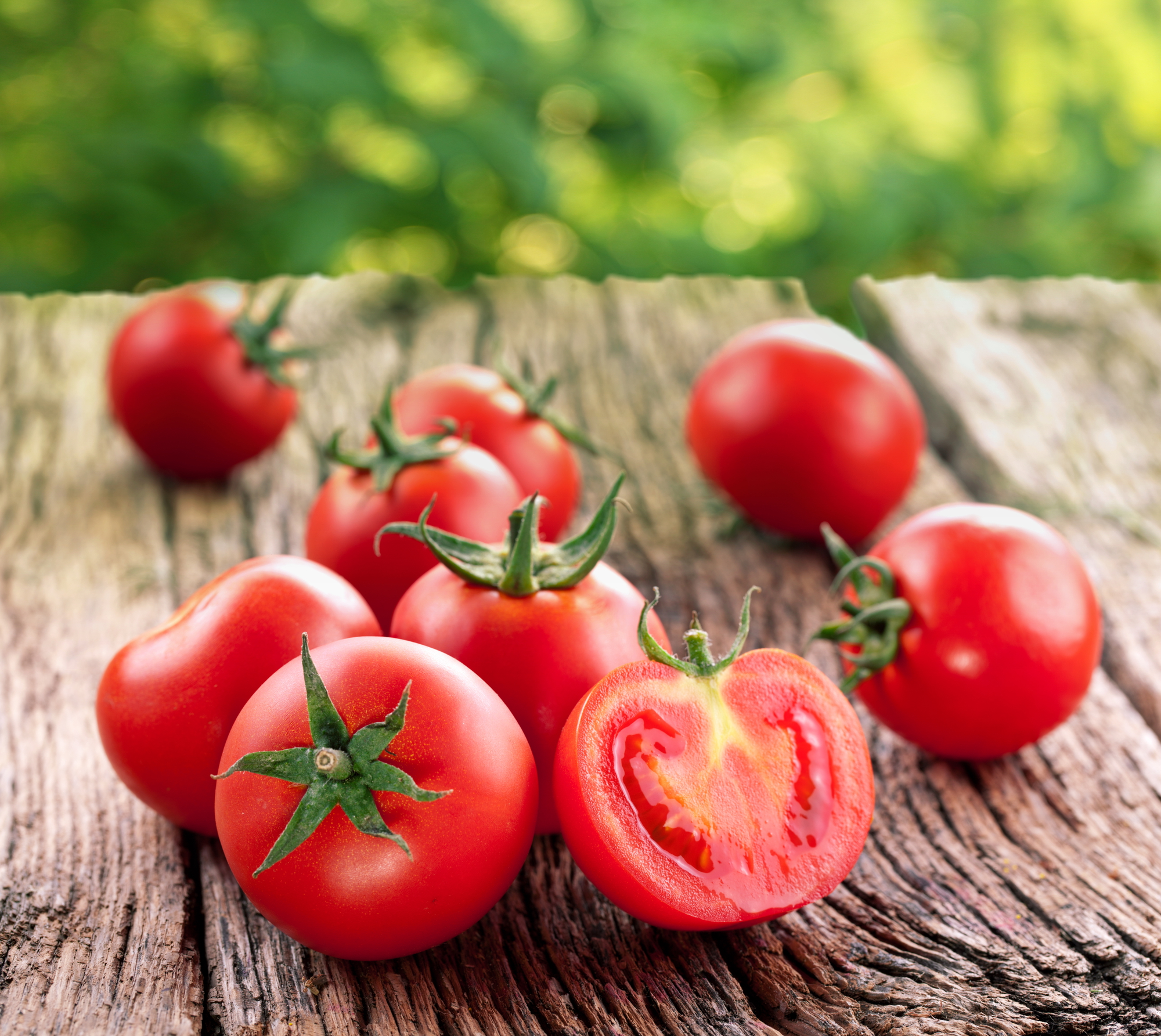 A group of ripe red tomatoes, including cherry tomatoes, sit on a rustic wooden surface with a blurred green foliage background. One tomato is cut in half, revealing its juicy interior.