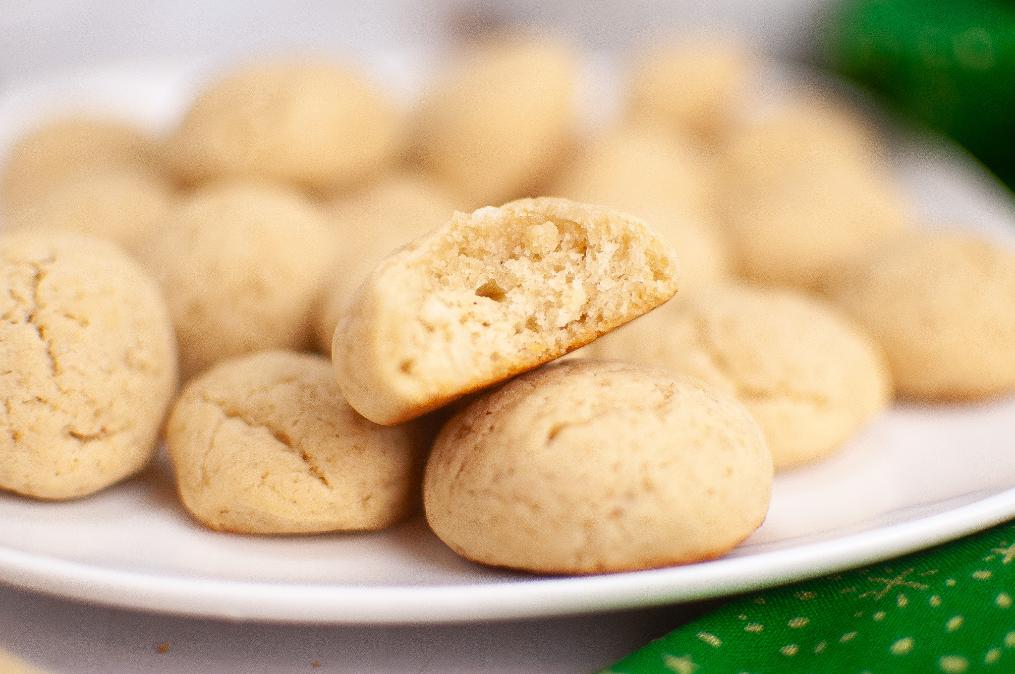 A plate full of soft, round Easy Old Fashioned Sour Cream Cookies on a white dish. One cookie is broken in half, revealing its soft, crumbly texture. The background is slightly out of focus, and a green cloth is partially visible.