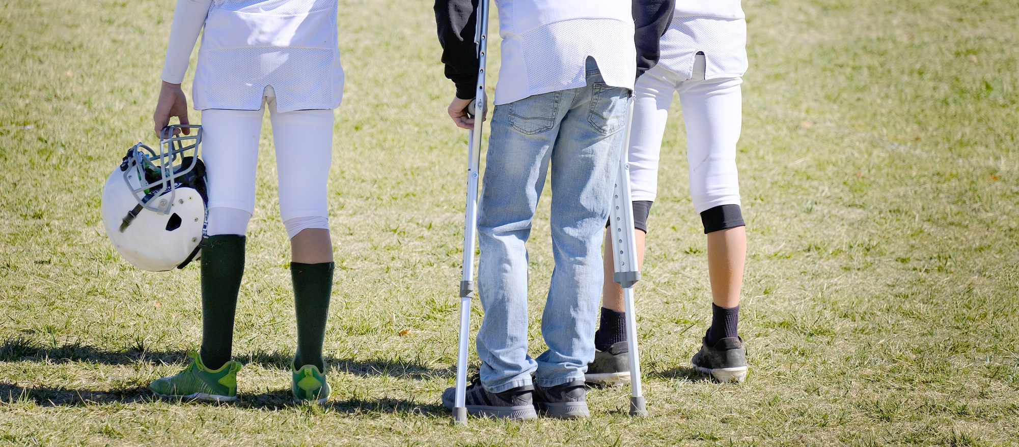 Three youth athletes stand in a field. Two wear football gear, one holding a helmet. The third, in jeans and a hoodie, uses crutches. Their backs are to the camera, and the sky is clear.