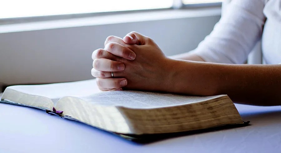 Hands together in prayer, laying on an open Bible.