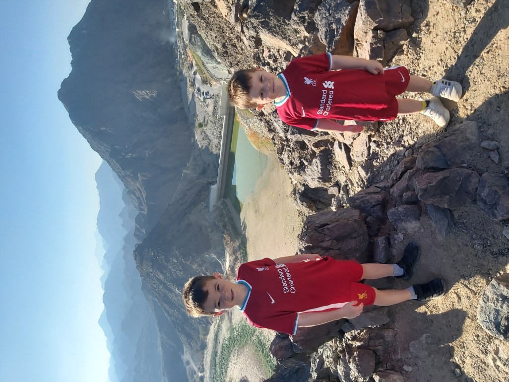 2 boys in Liverpool football kits at the top of a mountain on a family hike in UAE.