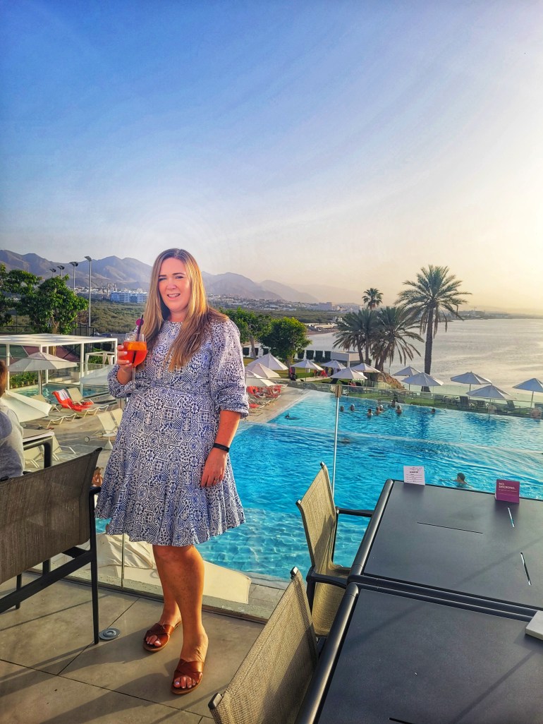 Woman holding an aperol spritz wearing a blue and white dress with a pool and sunset in the background. 