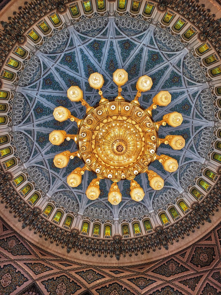 Looking up at a golden chandelier on the ceiling surrounded by gold and blue design. 