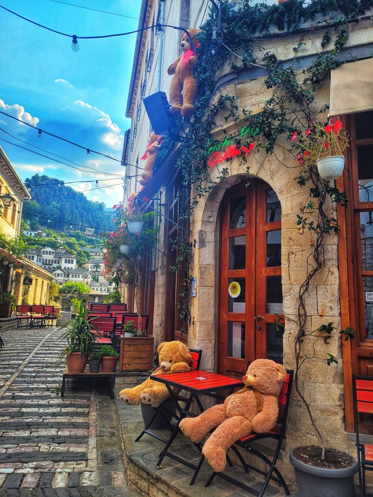 2 large teddy bears sitting at a table outside a traditional building on cobbled streets.