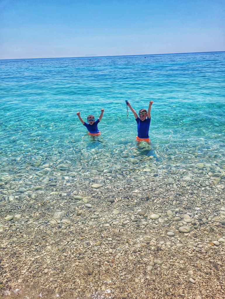 2 boys jumping in the clear turquoise sea with arms in their air. Dhermi has the most beautiful beaches for a holiday to Albania. 