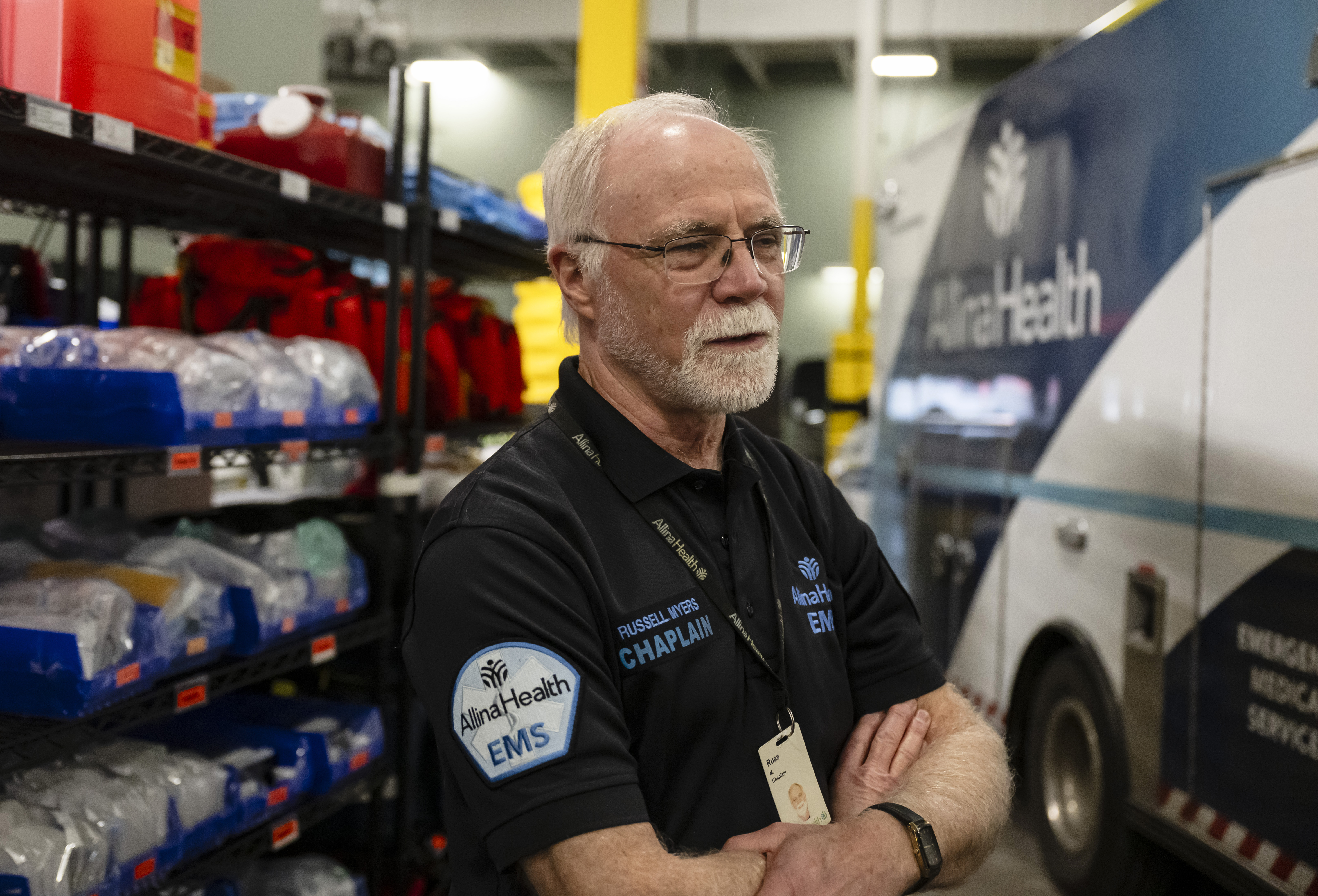 Russell Myers, wearing a shirt with 'CHAPLAIN' on it, crosses his arms as he stands near an ambulance.