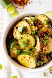 Sliced cucumbers in chili dressing sprinkled with sesame seeds and diced green onions in a white bowl.