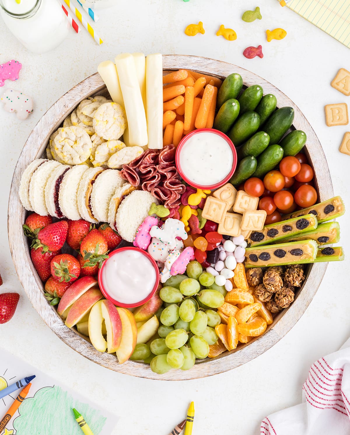 An overhead photo of a round snack board filled with fruits and veggies, sandwiches, cheese, and cookies.