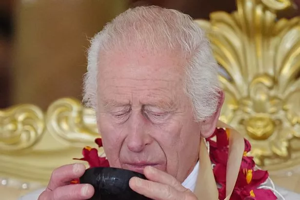 A photo of King Charles III drinking kava during a farewell ceremony at Siumu Village on the final day of the royal visit to Australia and Samoa. He is wearing a red scarf made of flowers and is wearing a grey suit