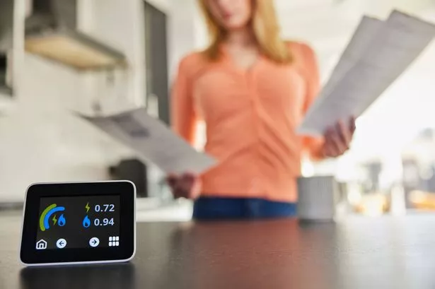 Close Up Of Smart Energy Meter In Kitchen Measuring Electricity And Gas Use With Woman Looking At Bills