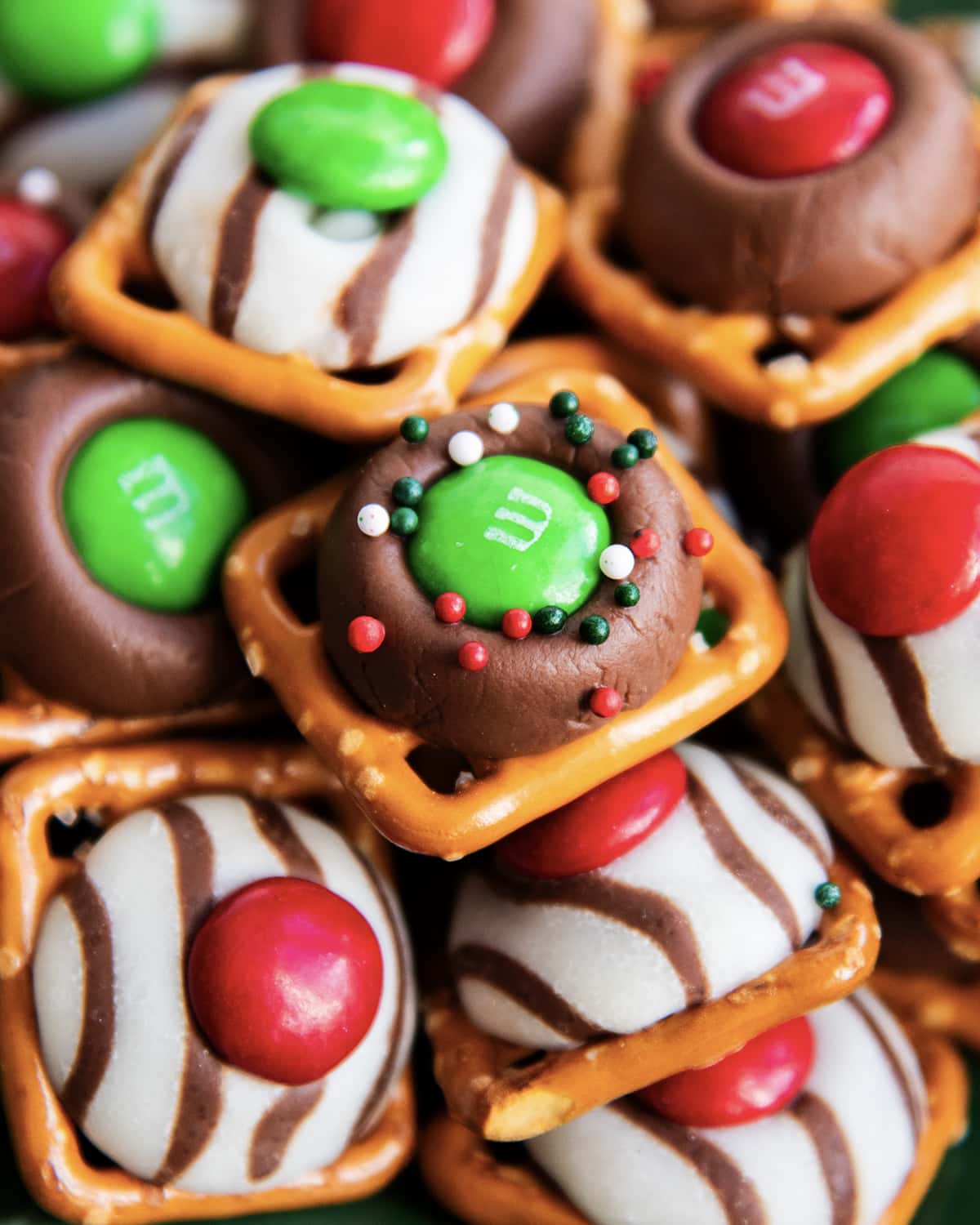 A close up of a pile of Christmas pretzel treats, with square snap pretzels, and topped with a green m&m and sprinkles.