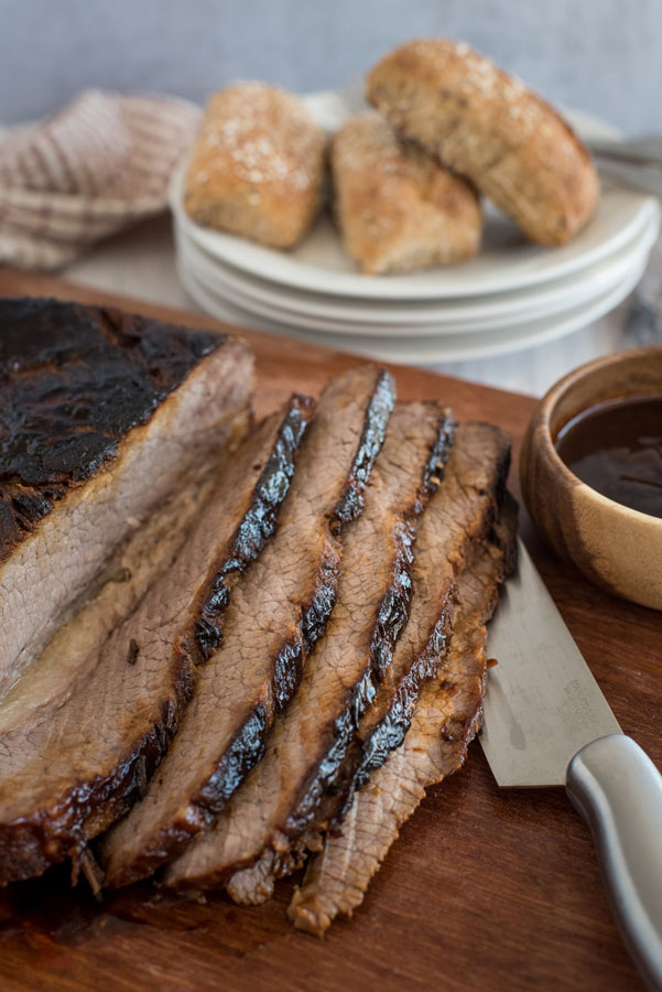 BBQ beef brisket cooked in an Instant Pot sliced on a cutting board with rolls in the back ground.