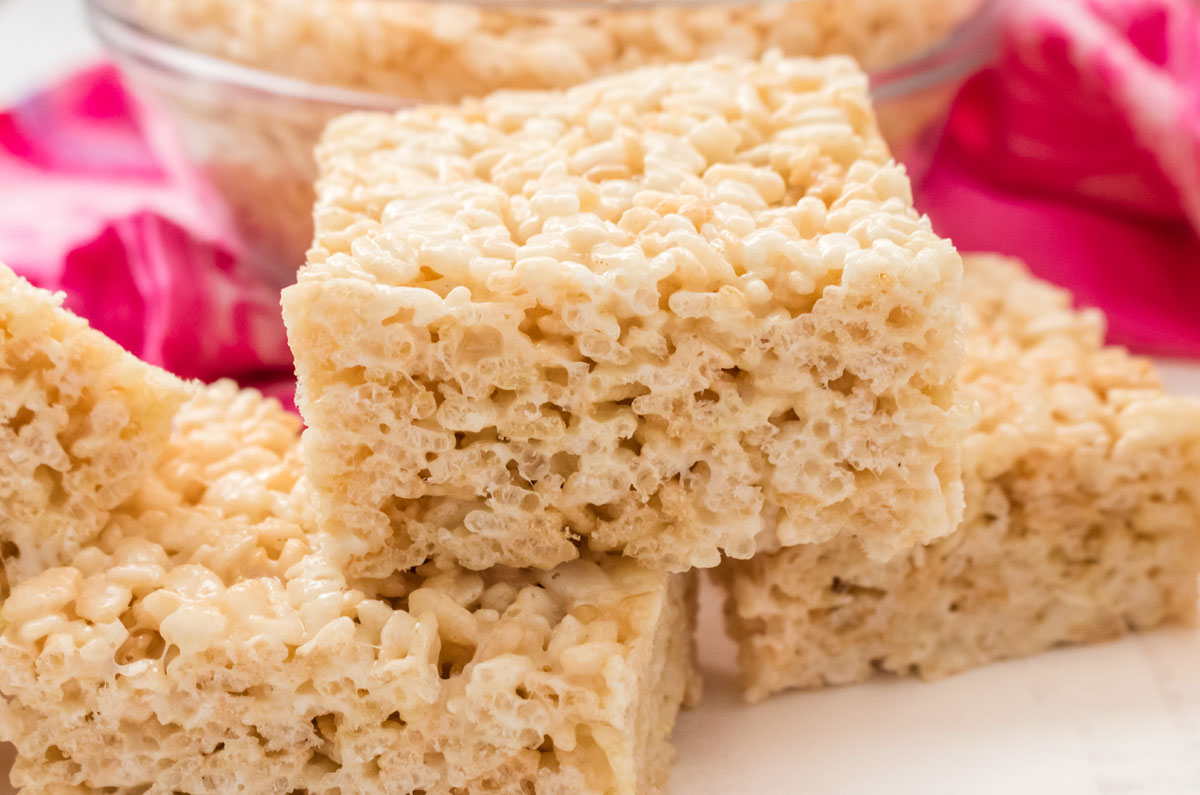 Three Rice Krispie Treats sitting on a white table in front of a glass bowl filled with Rice Krispie Cereal.