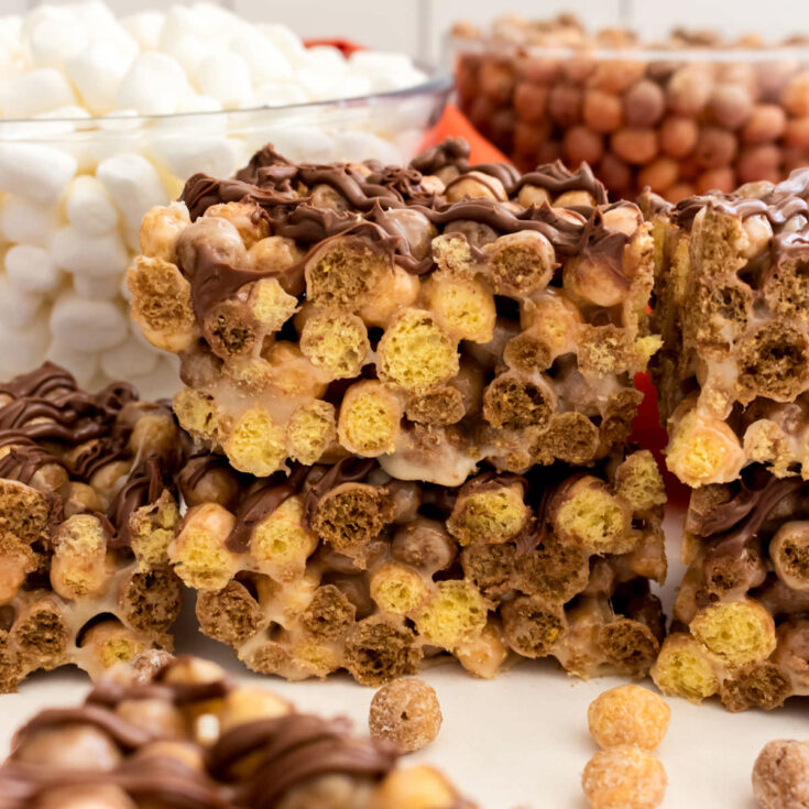 A batch of Reese's Puffs Marshmallow Treats sitting on a white table in front of clear bowls filled with marshmallows and cereal.