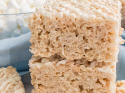 A stack of two White Chocolate Rice Krispie Treats sitting on a white table surrounded by glass bowls filled with Mini Marshmallows and Rice Krispies Cereal.