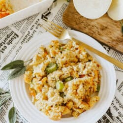 A plate of cornbread stuffing with celery is on a table covered with newspaper-patterned paper. A gold fork rests on the plate. Sage leaves are scattered nearby, alongside sliced onions and fresh celery on a wooden board.