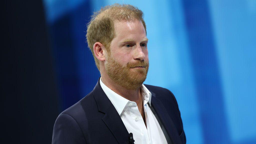 File photo of Prince Harry on a blue background, wearing a white open-neck shirt and a dark blazer.