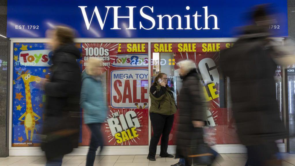 Blurred people walking past a WH Smith high street store which has large sale signs in the window