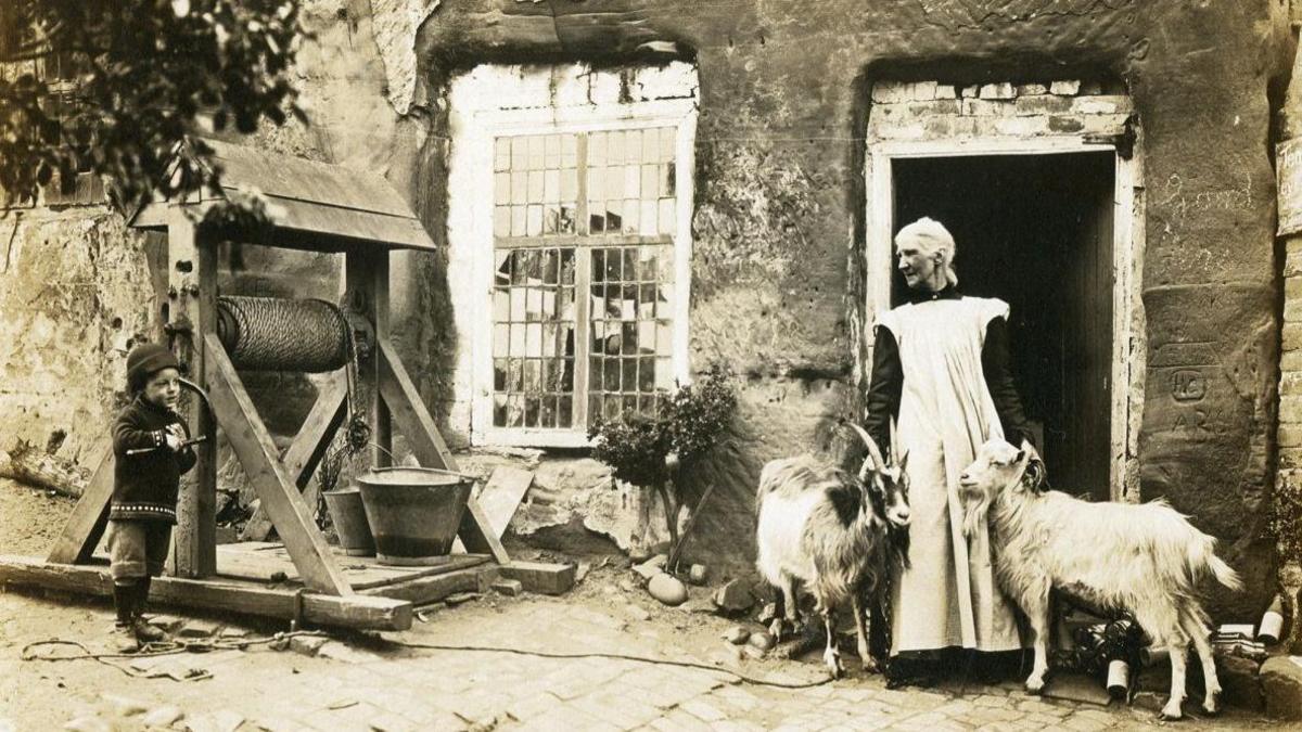 A woman in a white smock with black sleeves stands by a doorway with her hands each on the side of a goat. Beside them is a wooden well covering with a coiled up rope and a bucket. Beside the well is a small boy with a hat, jumper and trousers. 