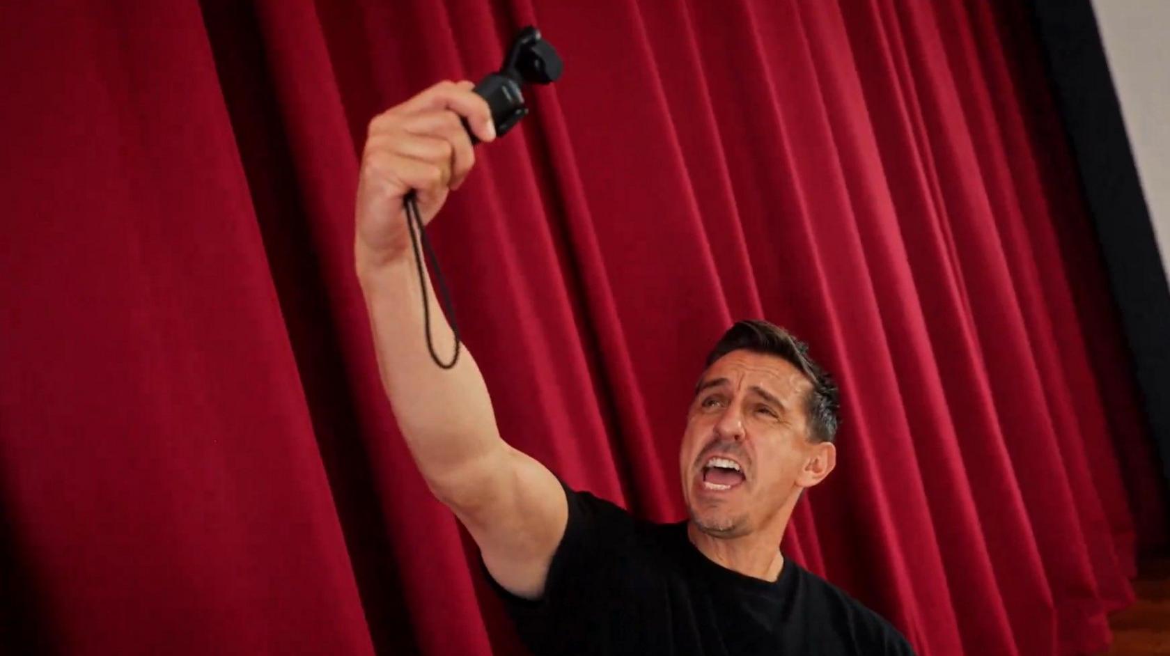 Gary Neville shouting into a GoPro camera on a stage, against the backdrop of a red stage curtain