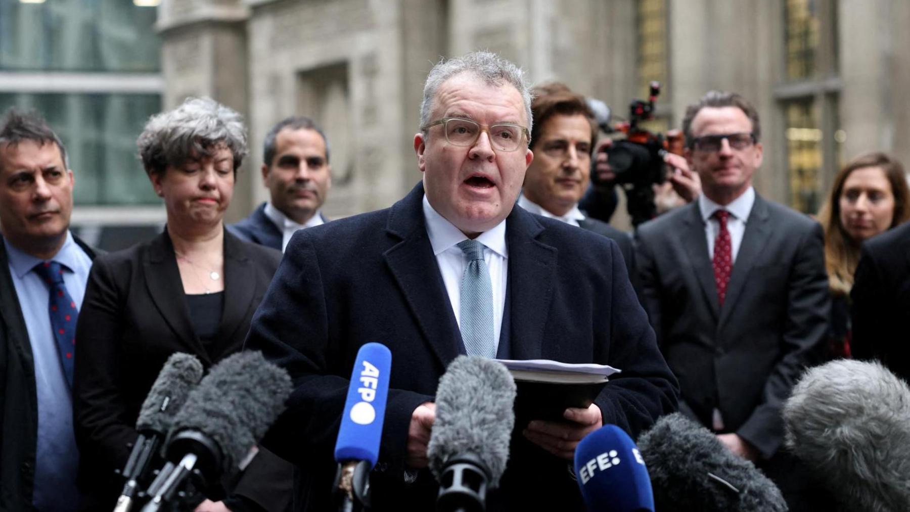 Tom Watson in dark suit speaking off notepad in front of a row of microphones. People in suits are standing behind him.