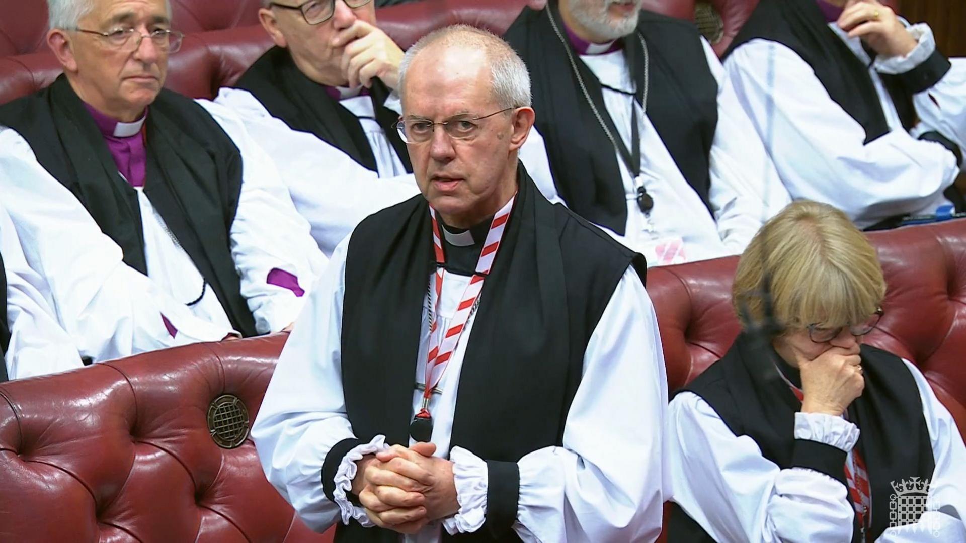 Archbishop Justin Welby stands, hands clasped, in the House of Lords chamber wearing black and white robes.