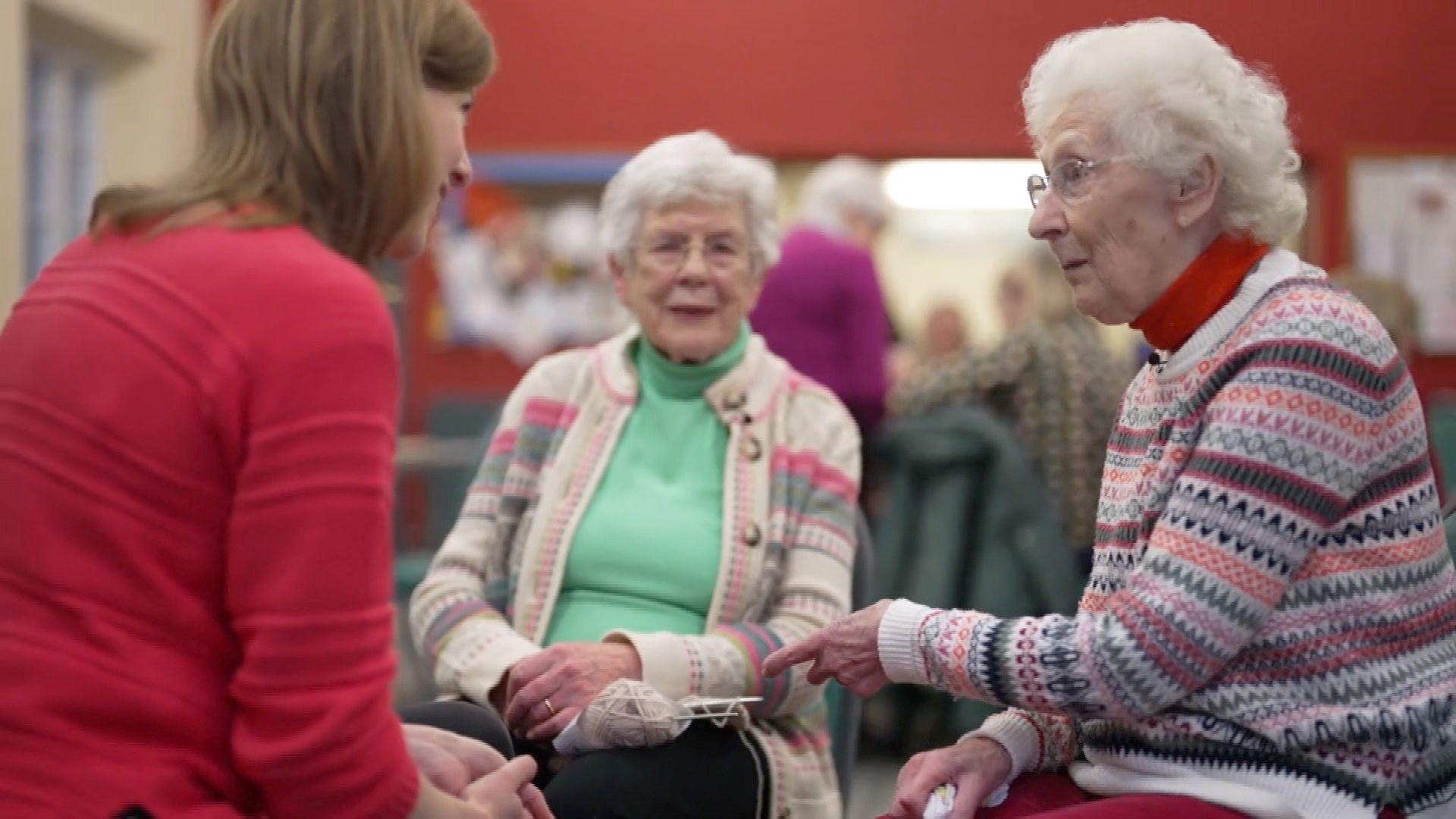 Friends Marjorie and Rosemary, both in their 90s, sit in a circle with BBC correspondent Colletta Smith. Colletta is wearing a red jumper, chatting to the two older women wearing knitted jumpers. One of the women is holding some knitting