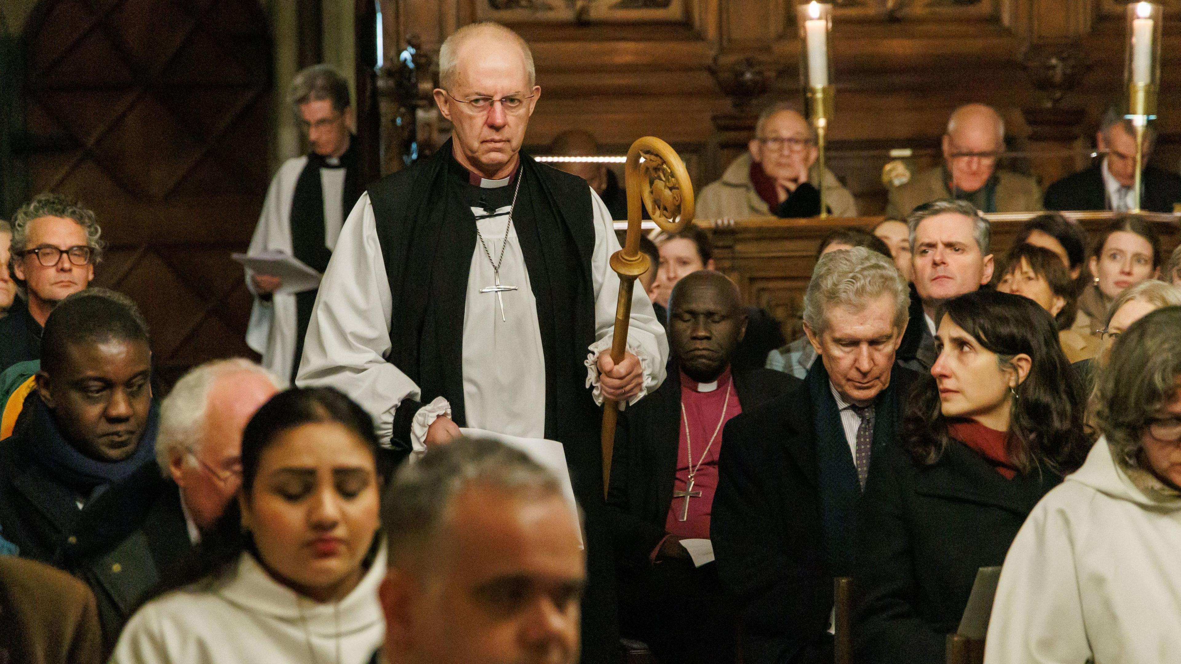 Justin Welby, arrives for a service of Evensong at Lambeth Palace Chapel. He carries the crozier and walks toward the altar, with the pews full of congregants on both sides of the aisle.