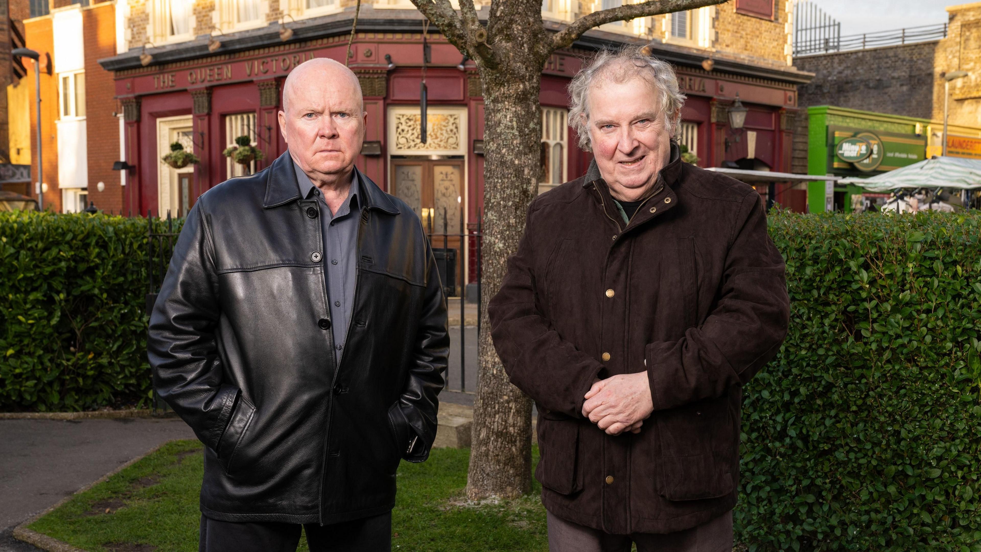 Paul Bradley as Nigel Bates (right) wearing a brown coat with this hands clasped in front of him, standing next to Steve McFadden as Phil Mitchell, who is wearing a black leather jacket with his hands in his pockets, in Eastenders.