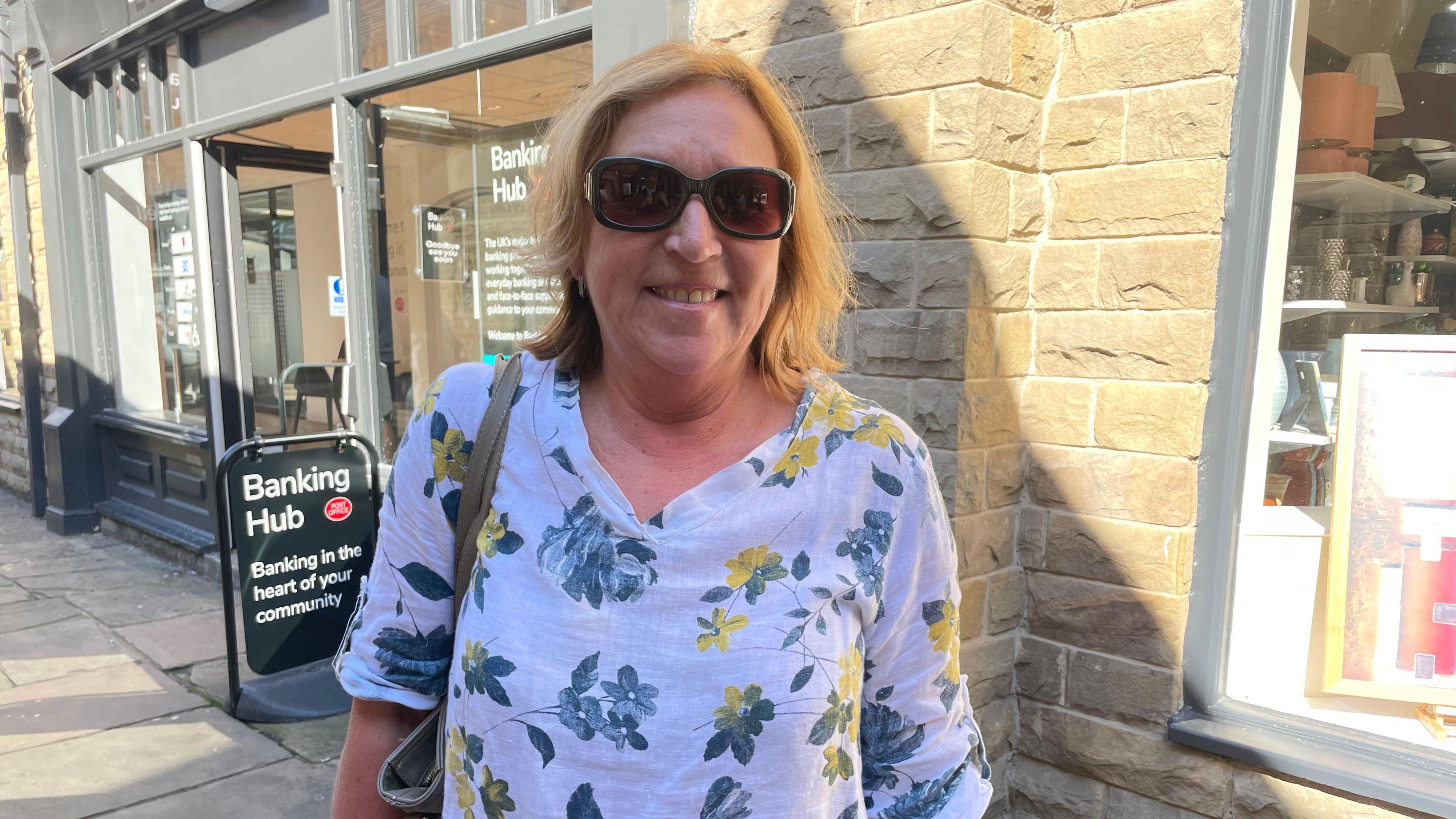 A smiling Lorraine Butterworth stands outside the banking hub. She wears a floral top and sunglasses, a bag is slung over her right shoulder.